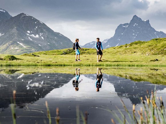 Ihr Hotel in St. Anton am Arlberg Zwei Wanderer auf einer grünen Wiese mit Bergreflexionen im Wasser