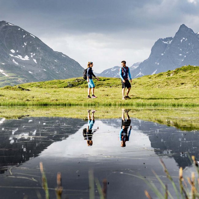 Ihr Hotel in St. Anton am Arlberg Zwei Wanderer auf einer grünen Wiese mit Bergreflexionen im Wasser