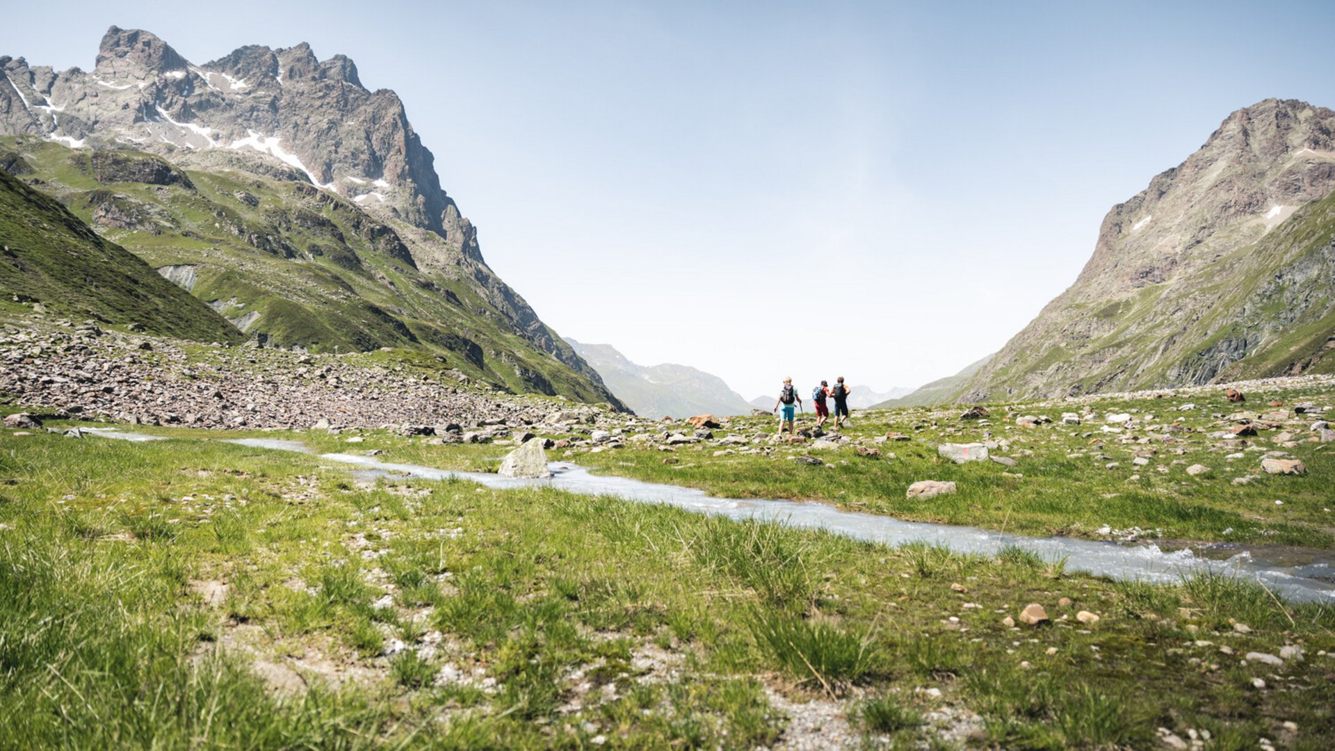 The pinnacle of mountain running Three hikers in a green valley with mountains and a stream under a blue sky