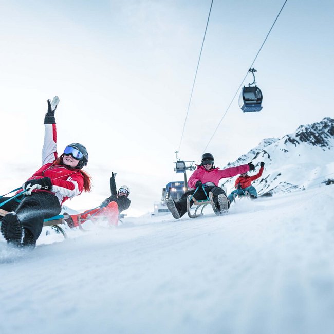 Reine Urlaubsfreude in Ihrem Hotel in St. Anton Vier Personen rodeln fröhlich einen schneebedeckten Hang hinunter mit Seilbahn im Hintergrund