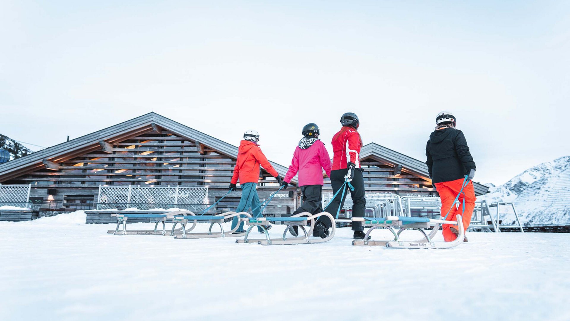 Reine Urlaubsfreude in Ihrem Hotel in St. Anton Vier Personen im Schnee mit Rodelschlitten vor Holzhütte und Bergen